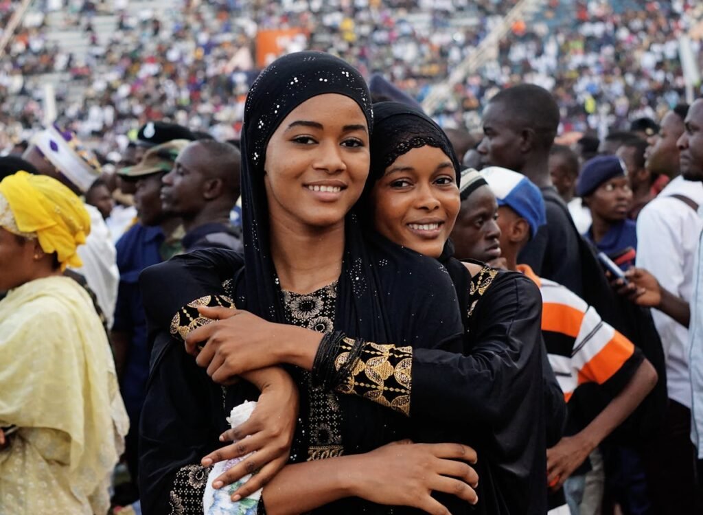 Positive young black female friends in traditional clothes and hijabs smiling and hugging while participating in event at stadium